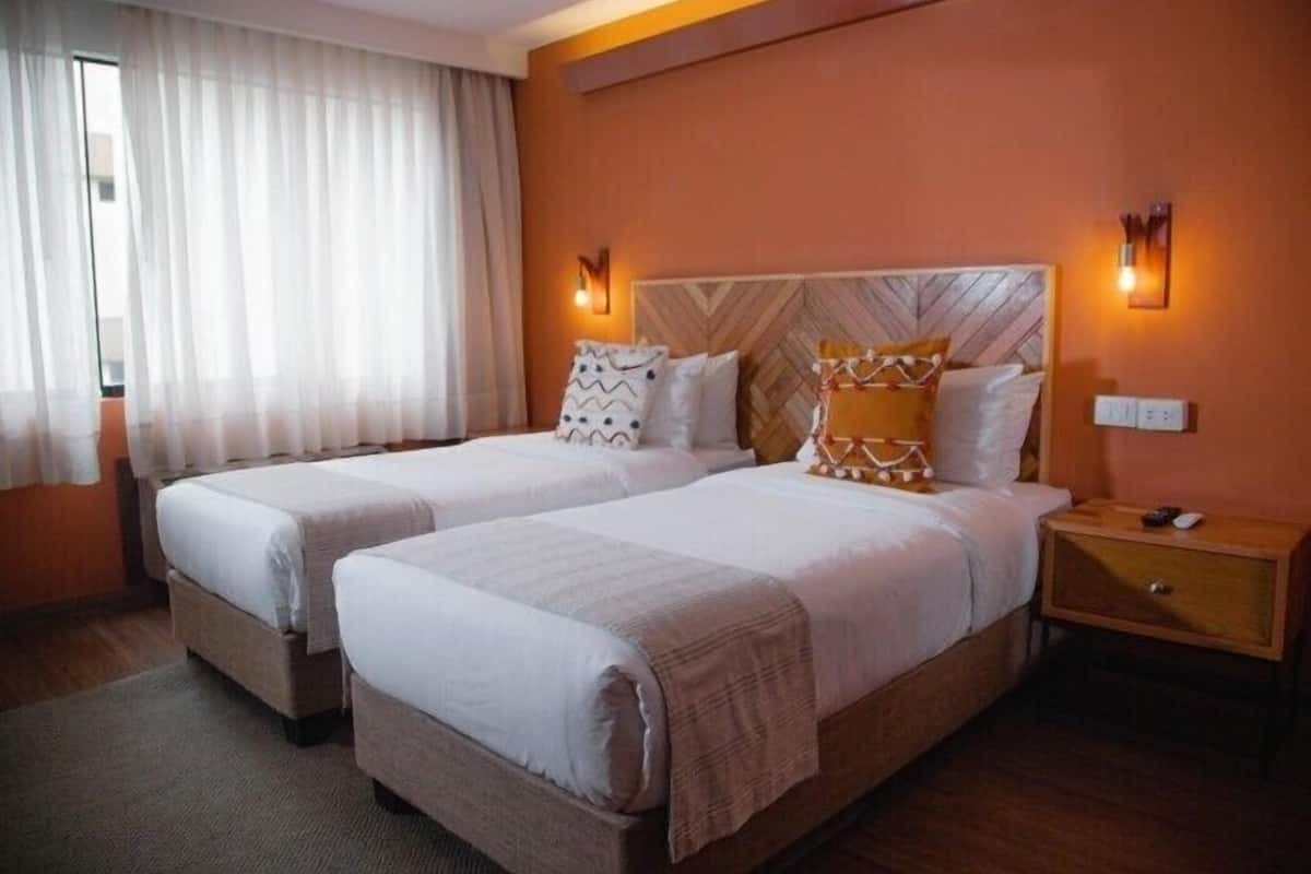 Habitación de hotel con dos camas, cabecera de madera, mesita de noche y ventana con cortinas traslúcidas.