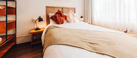 A neatly arranged hotel room with a bed, a wooden nightstand, a shelf with towels, a lamp, and a window with sheer curtains.