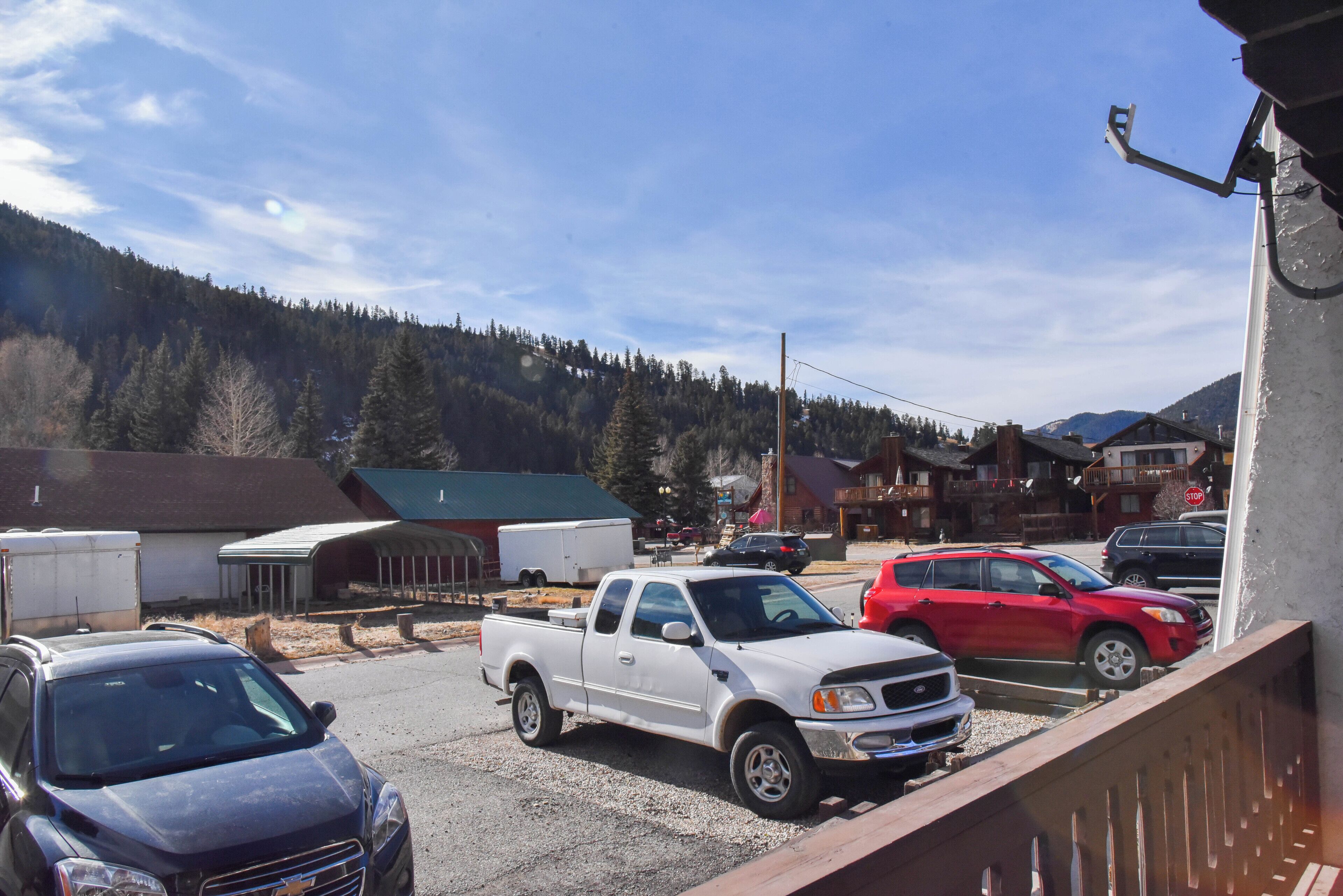 A parking lot with a white pickup truck, a red car, and a blue SUV. There are buildings and a mountain in the background.