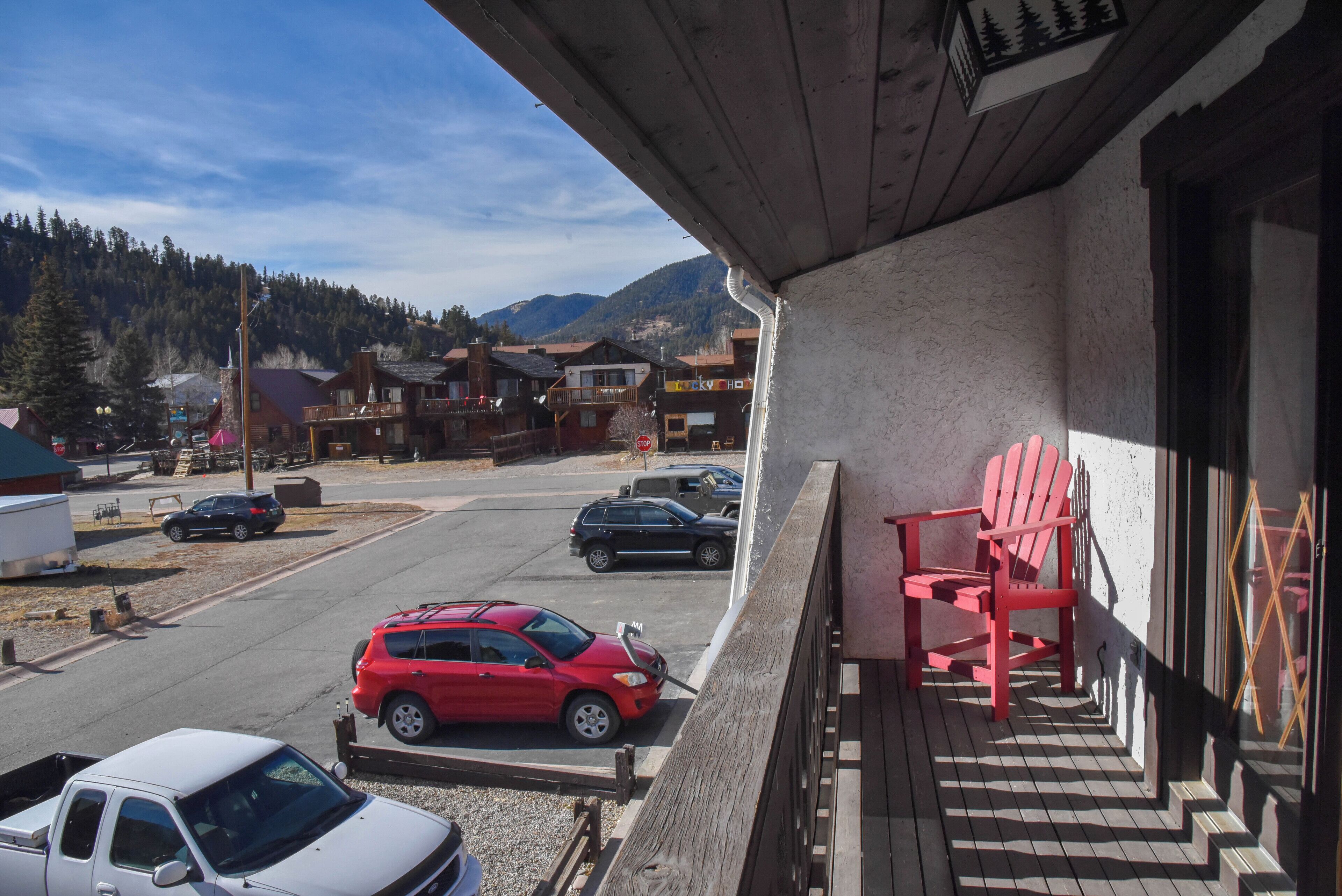 Un balcón con una silla Adirondack roja, vistas a un estacionamiento con varios autos y un paisaje montañoso.