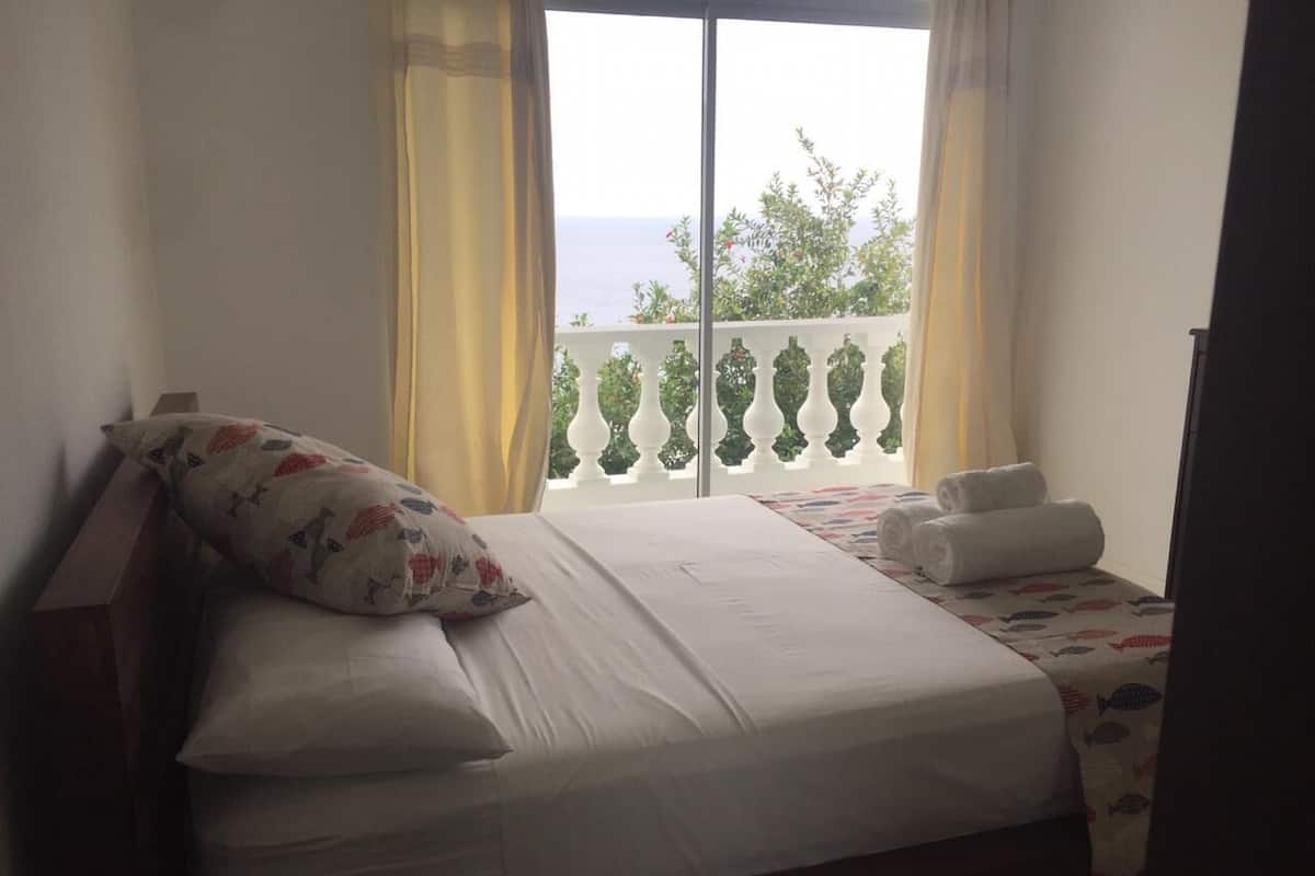 A hotel room with a bed, a window with curtains, a balcony with a railing, and a view of the sea.