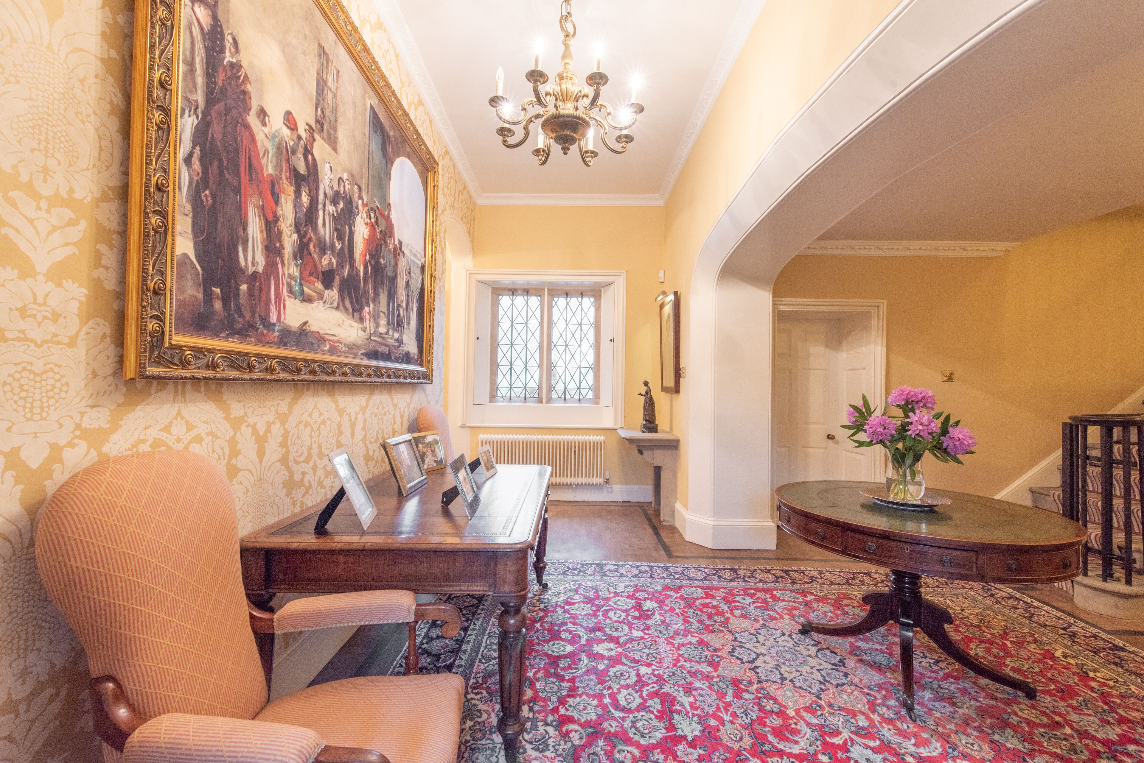 A dining area with a wooden table, chairs, a patterned rug, a chandelier, and a large framed painting on the wall.
