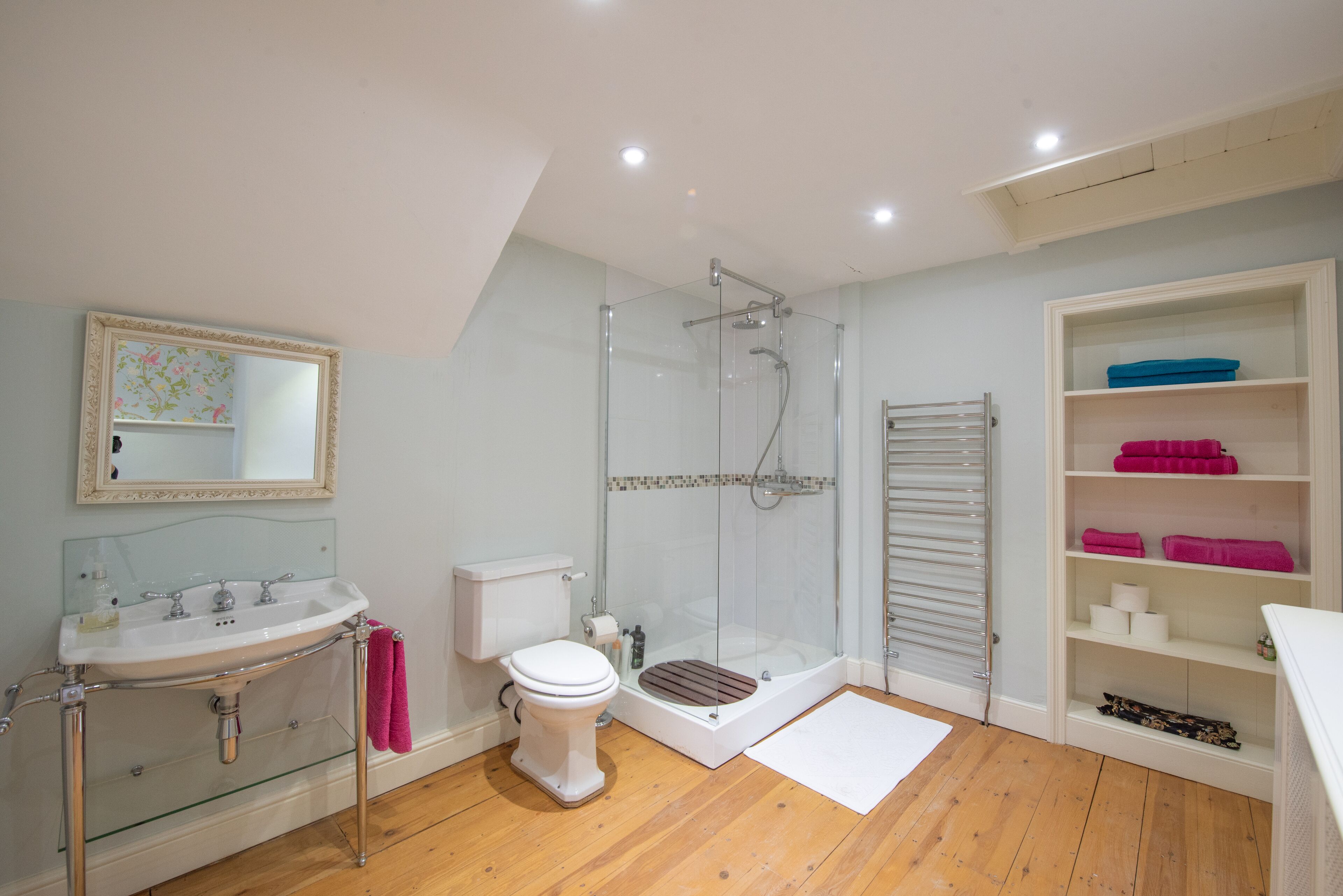 A modern bathroom with a glass shower enclosure, a white sink with a glass base, a toilet, and a built-in shelving unit with towels.