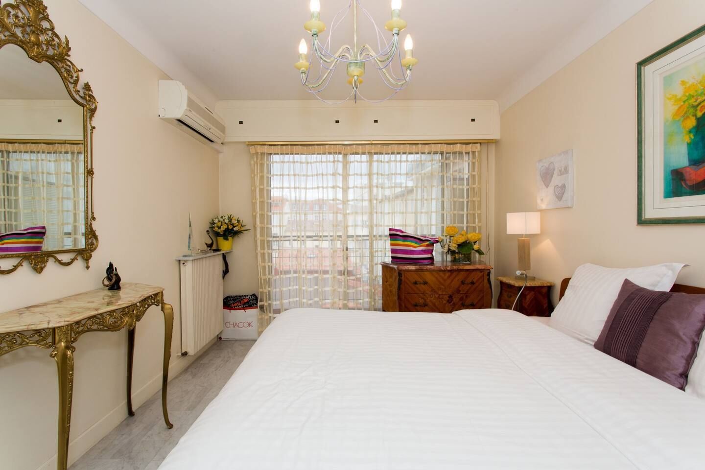 A hotel room with a large bed, a chandelier, a mirror, a dresser, a nightstand, and a window with curtains.