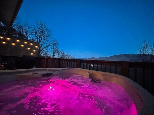 Un bain à remous avec un éclairage rose, entouré d’une terrasse en bois et avec une chaîne de montagnes en arrière-plan.