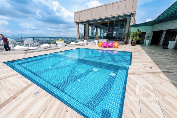 A rooftop pool area with a clear view of the city skyline, a modern building with glass facade, and a seating area with colorful chairs.