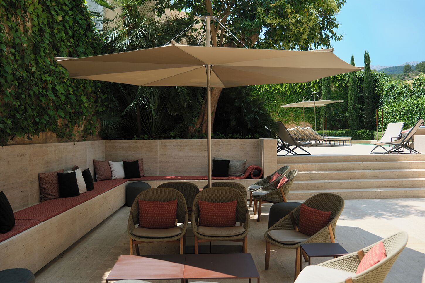 A sunlit outdoor seating area with wicker chairs and a large umbrella, surrounded by lush greenery and a pool.