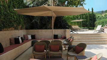 A sunlit outdoor seating area with wicker chairs and a large umbrella, surrounded by lush greenery and a pool.