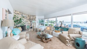 A spacious living room with a stone wall, large windows, and a view of the marina. - Beautiful Bayfront Retreat with Unmatched Water Views (Newport Beach)