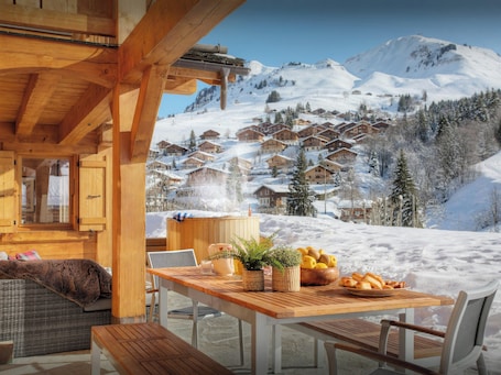 A wooden cabin with a table set for breakfast, overlooking a snowy mountain village.