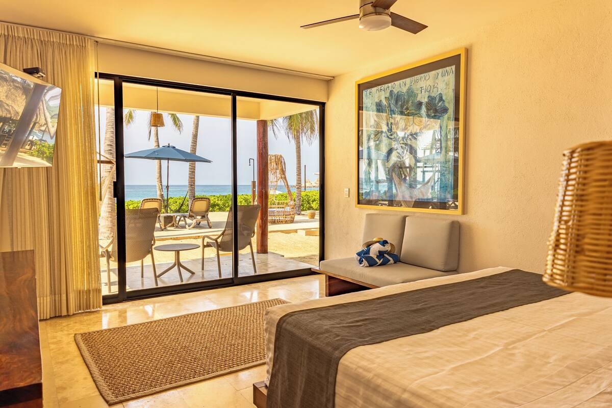 A hotel room with a bed, a large window with a view of the beach, a ceiling fan, and a painting on the wall.