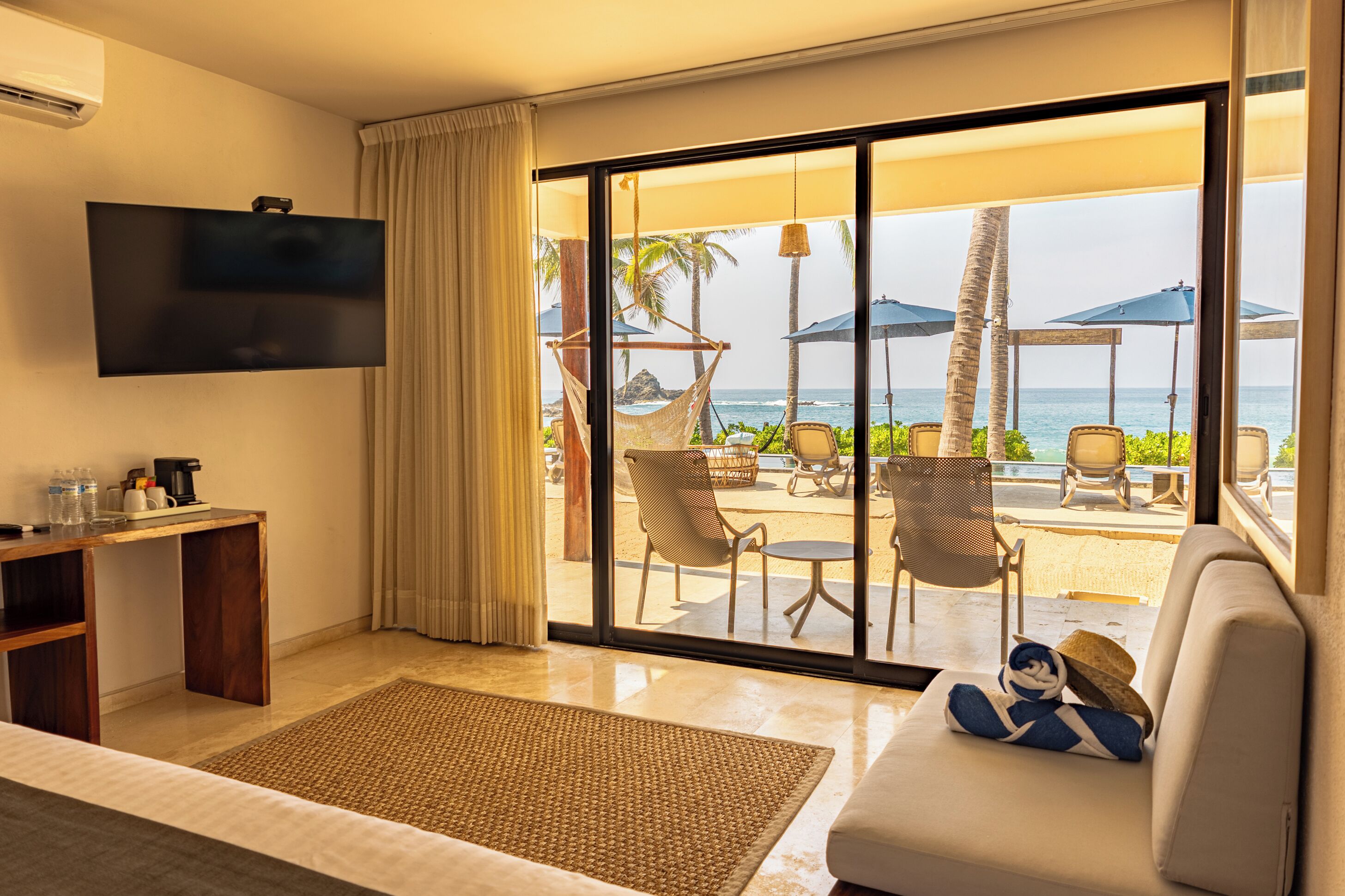 Ein Hotelzimmer mit Fernseher, einer Couch, einer Essecke mit Stühlen und Sonnenschirmen sowie Blick auf den Ozean.