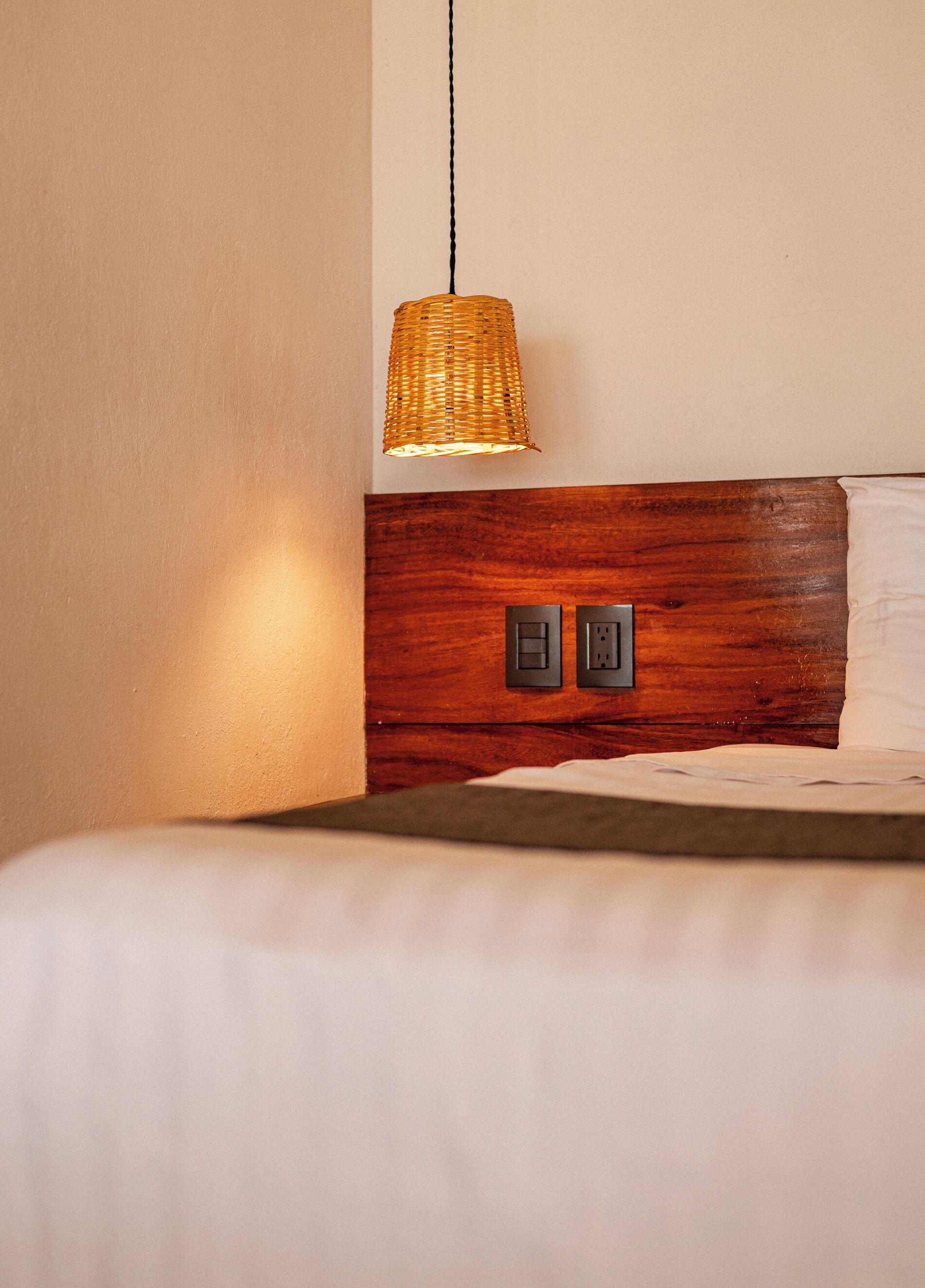 A bed with a wooden headboard, a woven light fixture, and two electrical outlets on the wall.