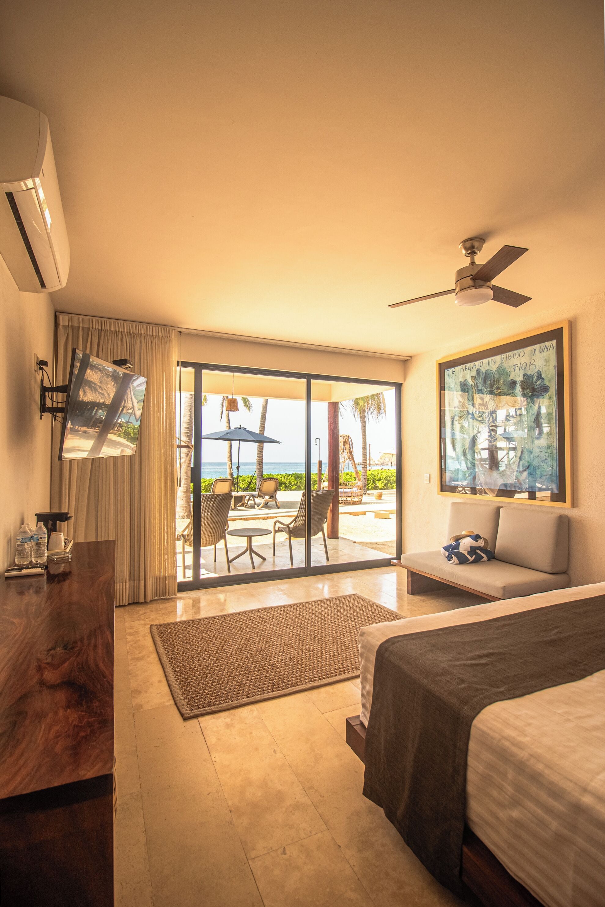 A hotel room with a bed, a desk, a flat-screen TV, a ceiling fan, and a sliding glass door leading to a balcony with outdoor furniture.