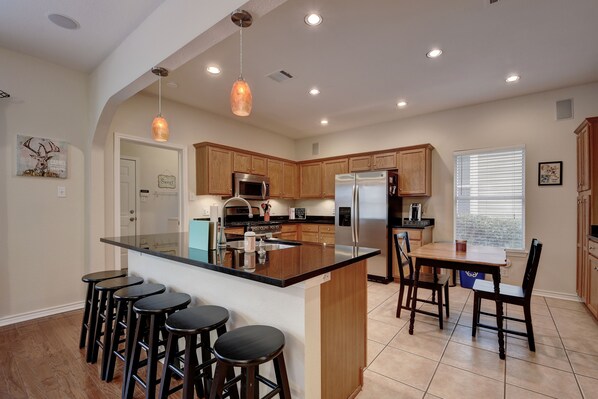 A modern kitchen with a central island, bar stools, wooden cabinets, stainless steel appliances, and pendant lighting.
