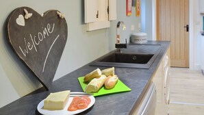 Une cuisine avec un panneau d’accueil en forme de cœur, un évier, une planche à découper avec du pain, une assiette avec du fromage et des tomates, et une porte en arrière-plan.