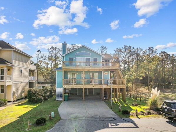 A two-story house with a balcony, a front porch, and a driveway leading to a parked SUV. - 1764 - Sunny Side Up - Oceanside Corolla Retreat | Pool & Hot Tub (Corolla)