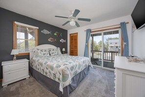 Un dormitorio con cama, mesita de noche, ventilador de techo, ventana con persianas, puerta corrediza de vidrio y vista al exterior.