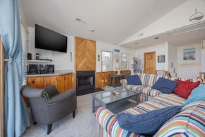A living room with a TV, speakers, a fireplace, striped sofas, and a bookshelf. - 1764 - Sunny Side Up - Oceanside Corolla Retreat | Pool & Hot Tub (Corolla)