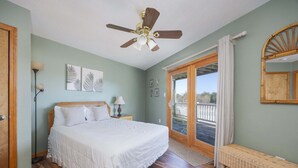 Une chambre à coucher avec un lit, un ventilateur de plafond, un plancher en bois et une vue sur l’extérieur, visible à travers une porte vitrée.