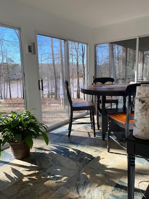 A sunlit room with a dining table and chairs, large windows offering a view of trees and a body of water.