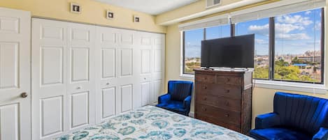 Un dormitorio con una cama, un aparador, dos sillas, una ventana con vistas y una puerta cerrada.