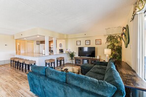 A living room with a large sofa, a TV, and a kitchen area with a bar. - XPLORIE ACTIVITIES & LINENS INCLUDED*! 3BR/2BA Capri. Spectacular Bay View! (Ocean City)