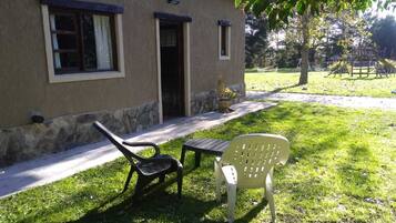 A building with a stone foundation and a beige exterior. There are two doors, one open and one closed. In front of the building, there is a small table and two chairs, one black and one white.