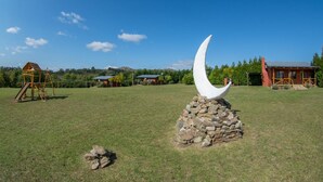 A grassy field with a large white crescent sculpture on a stone base, surrounded by buildings and trees. - Cabañas La Toscana (Tandil)