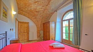 Une chambre à coucher avec une couvre-lit rouge, une armoire en bois, une grande fenêtre en arc de cercle et un plafond en briques.