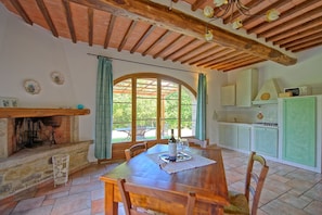 Un coin repas rustique avec une table et des chaises en bois, une cheminée et une vue sur une terrasse extérieure, accessible par une grande fenêtre en arc de cercle.