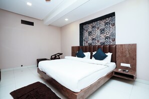 A modern bedroom with a large bed, a headboard with a patterned tapestry, a bedside table with a phone and remote, and a small rug on the floor. - The White Fresco (Udaipur)