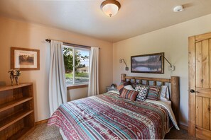 A bedroom with a bed, a window with curtains, a wooden door, a bedside table with a lamp, and a framed picture on the wall.