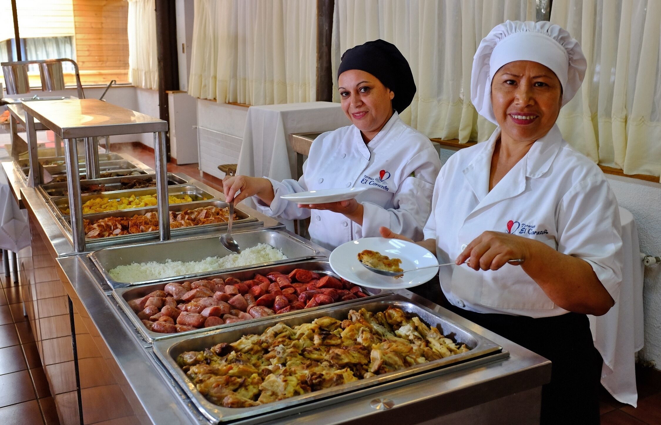 Un buffet allestito con diversi cibi e due membri dello staff che servono.
