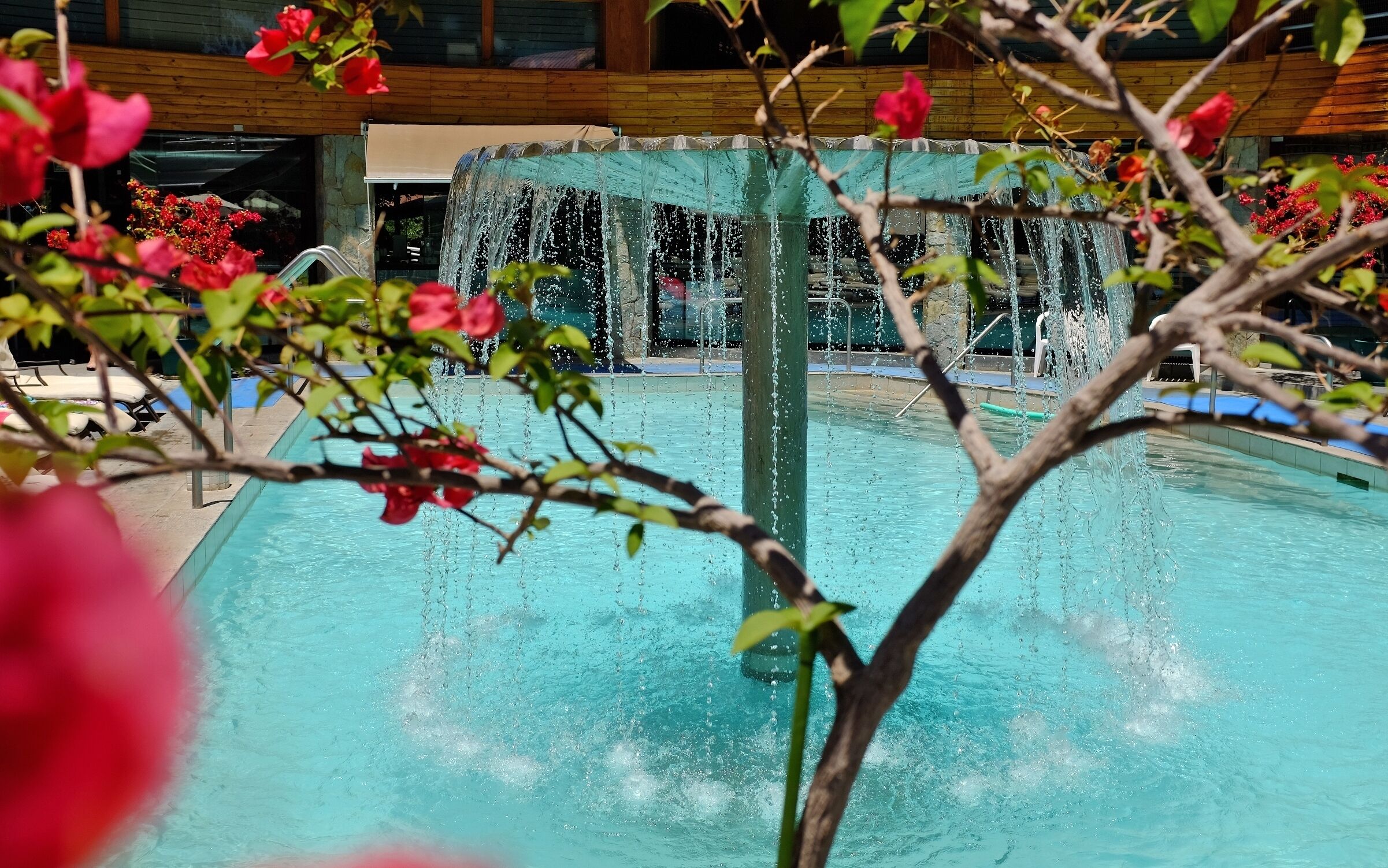 A pool with a central fountain, surrounded by lounge chairs and a wooden deck.