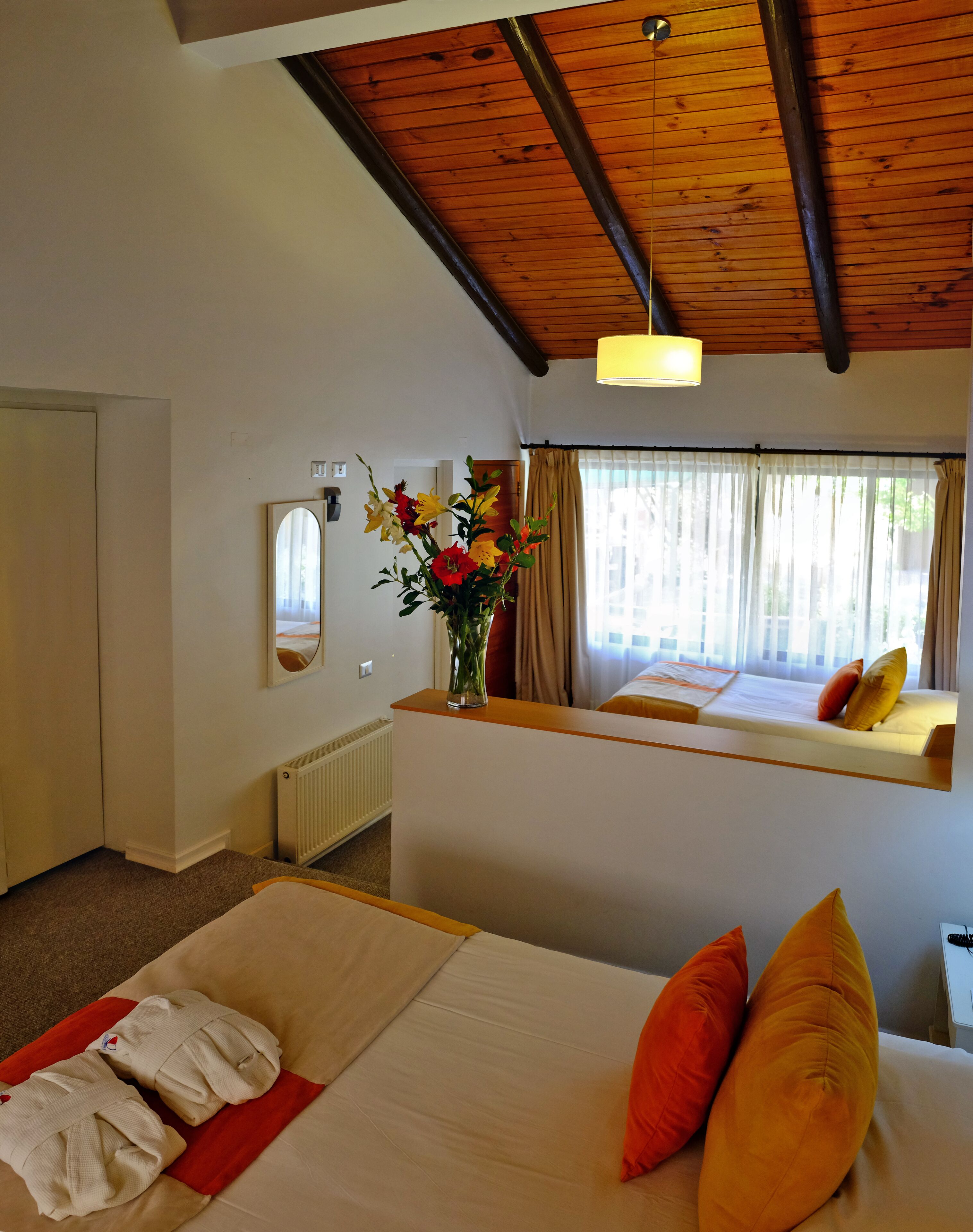Una habitación de hotel con cama, espejo, ventana con cortinas, un jarrón con flores y techo con vigas de madera.