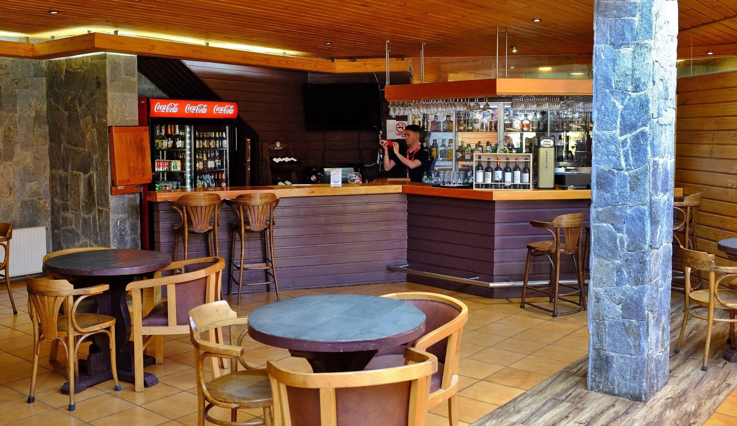 Een bar met een stenen pilaar, een houten toog en krukken. Op de toog staan flessen en glazen, en er is een bord met Coca-Cola.