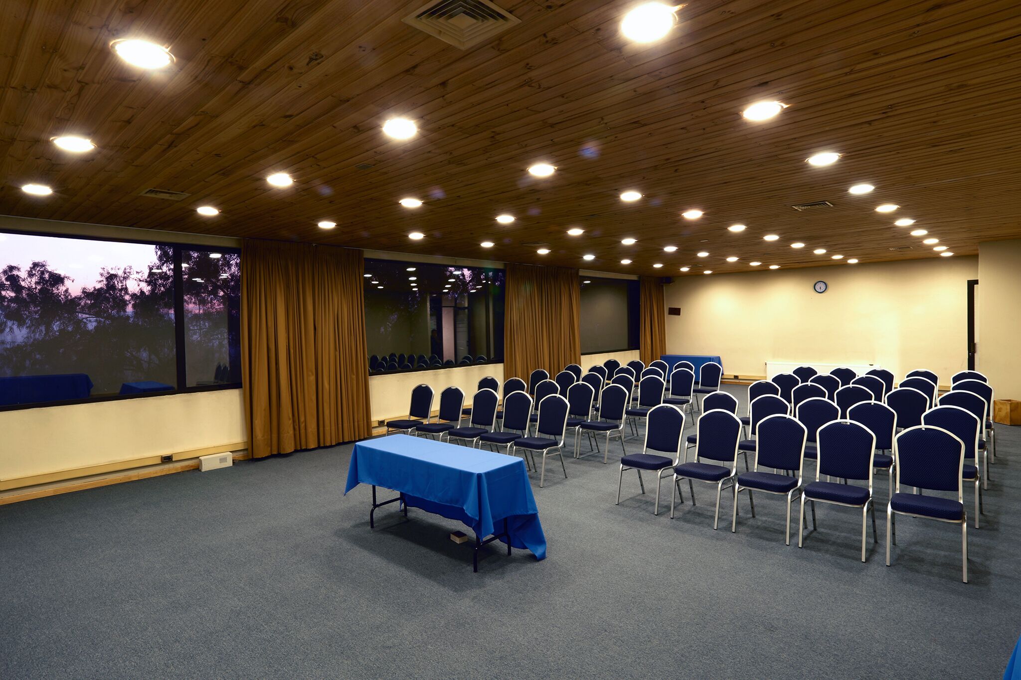Une salle de conférence avec une table bleue, des rangées de chaises, un plafond en bois et de grandes fenêtres.
