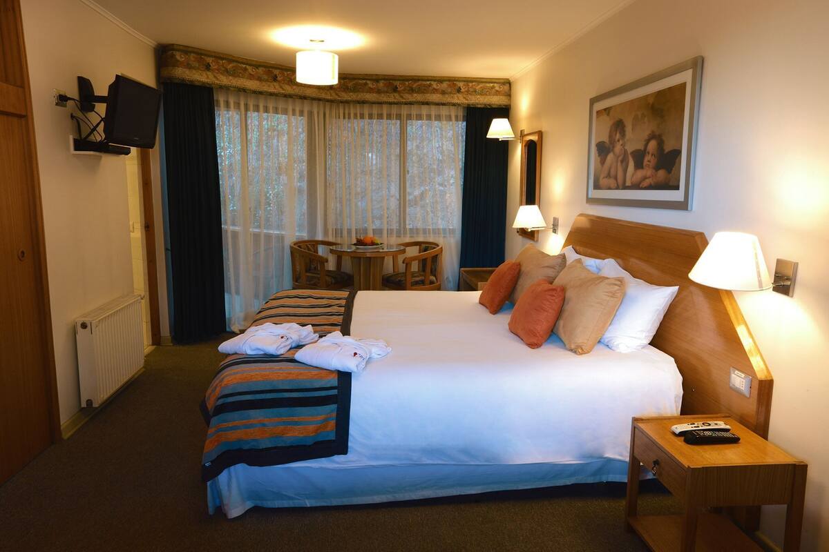 A hotel room with a bed, bedside table, lamp, TV, and a dining area with a table and chairs.