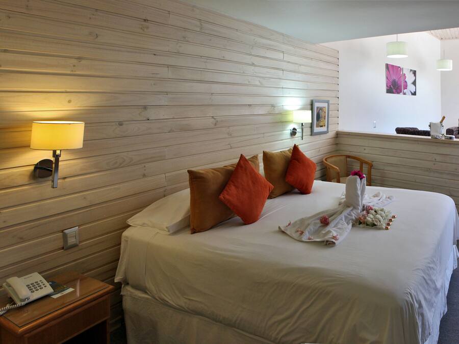 A hotel room with a wooden wall, a bed with white bedding and orange pillows, a bedside lamp, a telephone, and a small table with a lamp.