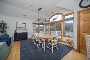 A dining area with a wooden table and white chairs, a chandelier, and large windows with a view of the outdoors.