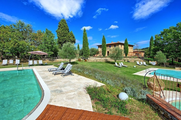 Zona de piscina con sillones de descanso, una terraza de piedra y un edificio de piedra al fondo.