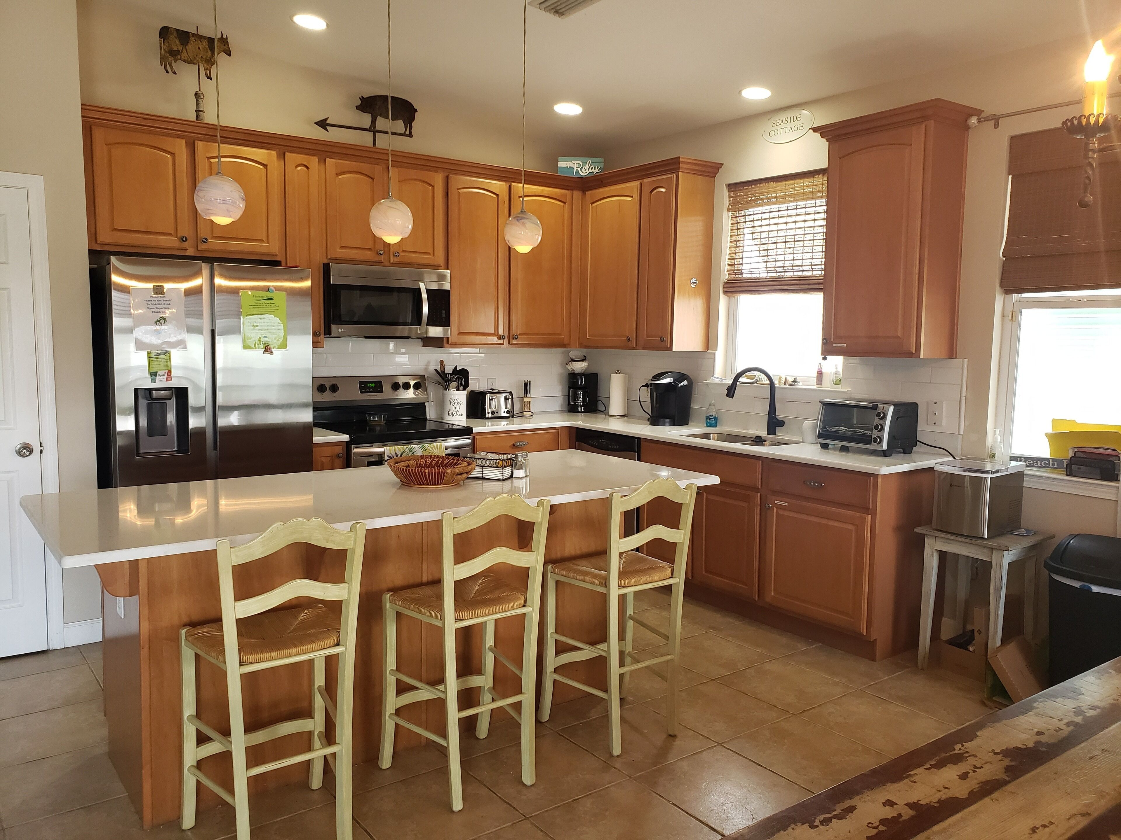 A kitchen with wooden cabinets, a stainless steel refrigerator, a microwave, a stove, and a sink. There are three bar stools and a large island with a bowl.