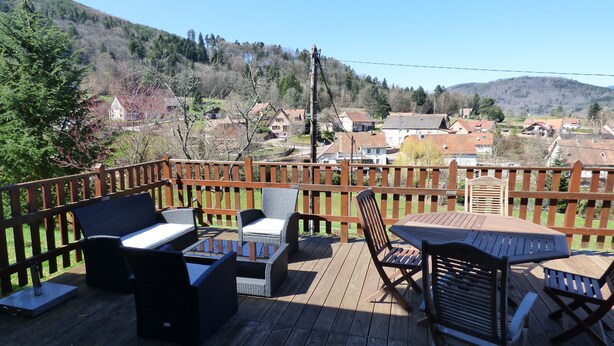 Eine Holzterrasse mit Gartenmöbeln, darunter Sofas und Stühle, mit Blick auf eine Wohngegend mit Häusern und Hügeln im Hintergrund.