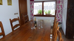 Ein Essbereich mit einem Holztisch, Stühlen und einer Champagnerflasche. Auf der Fensterbank stehen Blumentöpfe und es gibt floral bedruckte Vorhänge.
