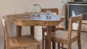 Uma sala de jantar com mesa e cadeiras de madeira, uma janela com persianas e uma cozinha ao fundo.