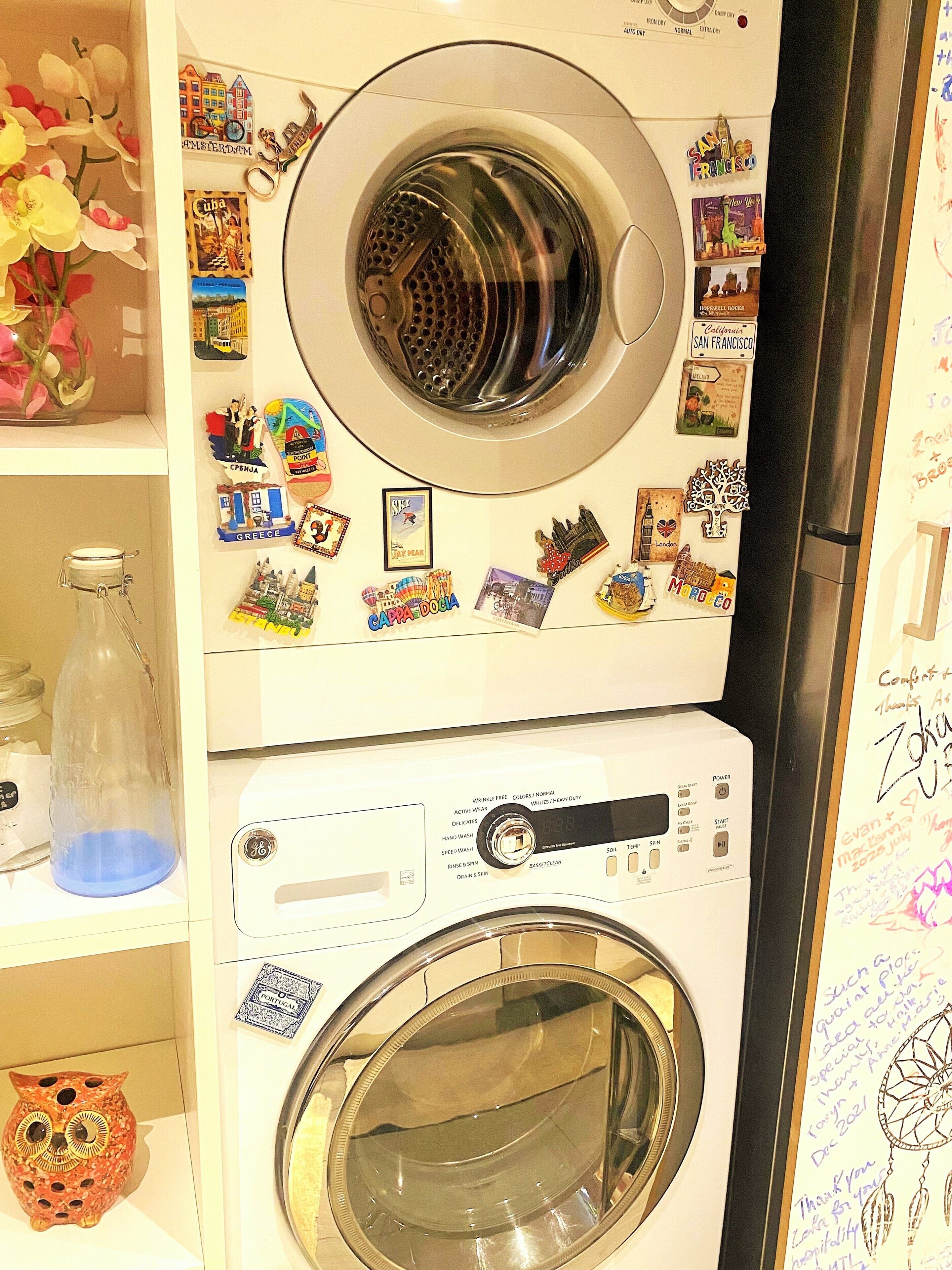 A front-loading washing machine with a control panel, surrounded by various travel-themed magnets and stickers.