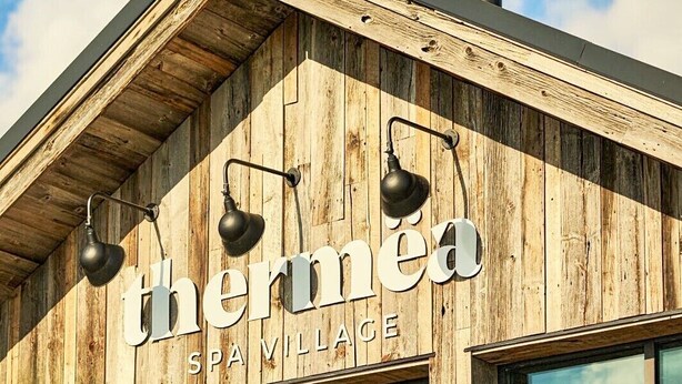 A building with a wooden facade, the name 'thermea SPA VILLAGE' prominently displayed, and three attached light fixtures.