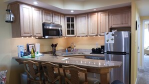 A kitchen with wooden cabinets, a stainless steel refrigerator, a microwave, and a sink. There are three wooden chairs at the island.