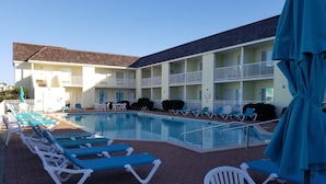 A pool area with lounge chairs and umbrellas, a building with multiple balconies, and a brick walkway.